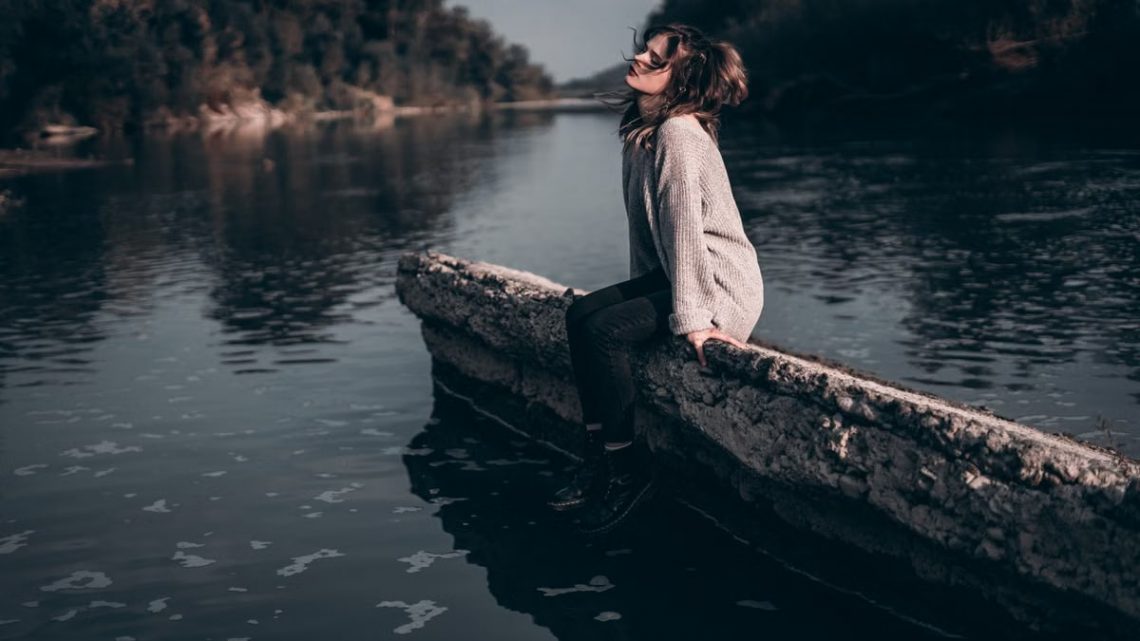 A woman enjoys a peaceful moment sitting on a riverside barrier during sunset.