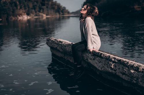 A woman enjoys a peaceful moment sitting on a riverside barrier during sunset.